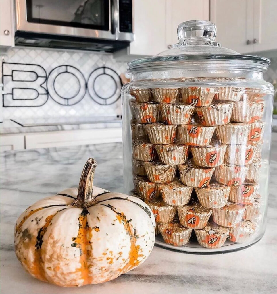 Large apothecary jar filled with wrapped candies beside a small white-and-orange pumpkin on the counter.