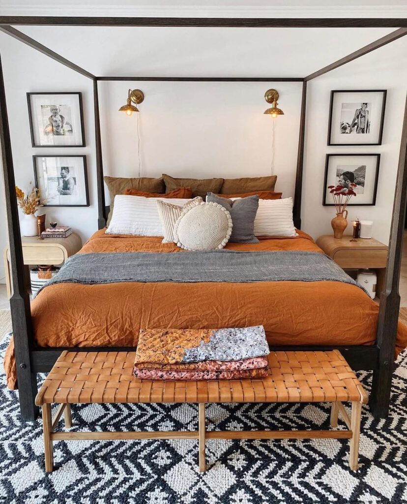 Modern canopy bed with rust duvet, brass sconces, striped pillows, woven bench and bold rug — modern fall bedroom ideas.