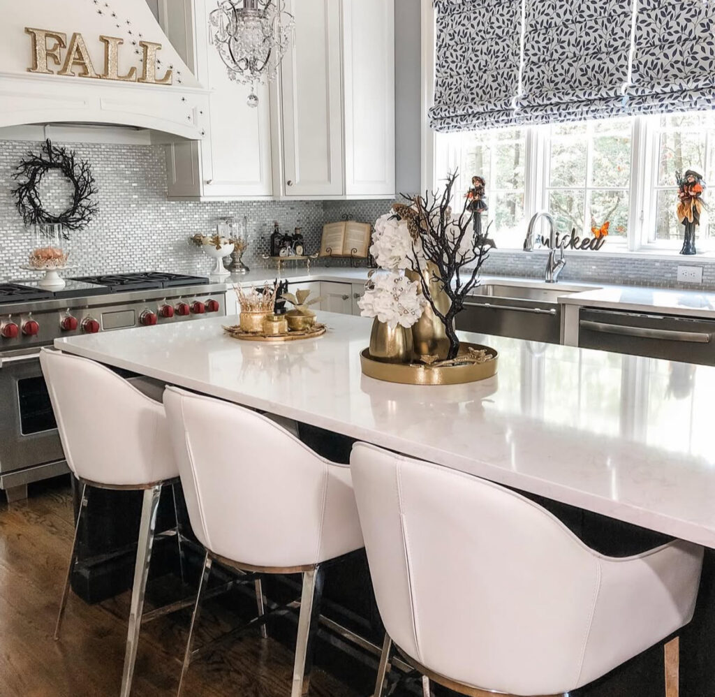 Glam white kitchen with a gold tray, black twig branches, metallic pumpkins, and a “wicked” sign at the window.