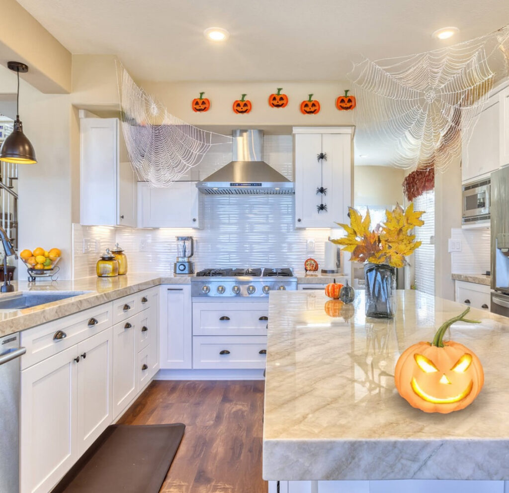 Bright kitchen with oversized faux spiderwebs, climbing black spiders, pumpkin faces above cabinets, and a carved jack-o’-lantern on the island.