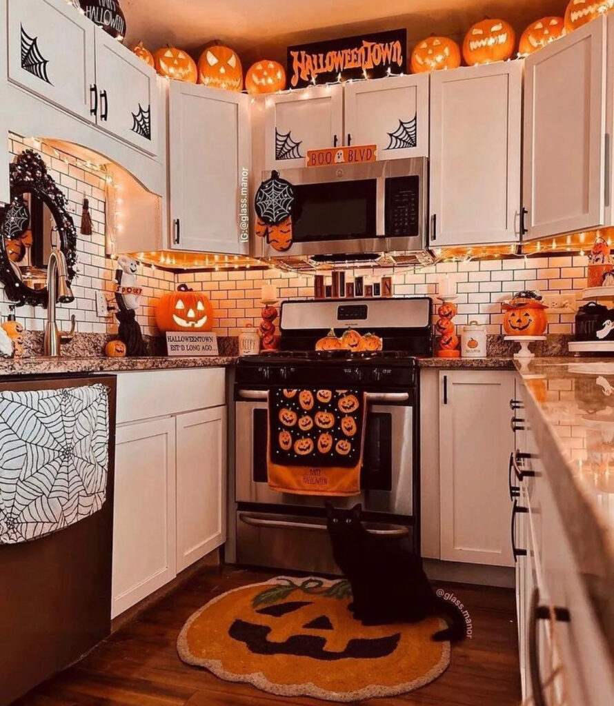 Cabinet tops lined with glowing jack-o’-lanterns and string lights; pumpkin rug and black cat below.