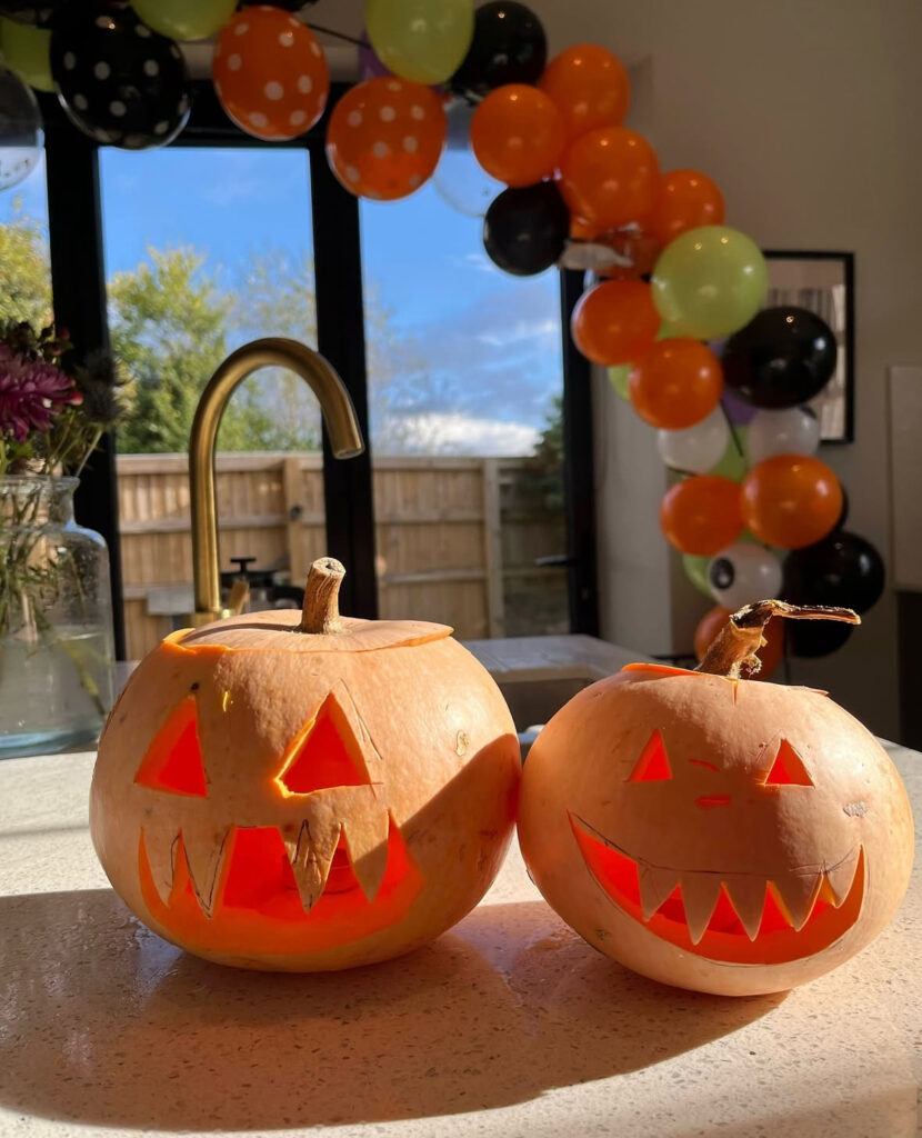 Two glowing jack-o’-lanterns on a kitchen island with an orange, green, and black balloon arch.