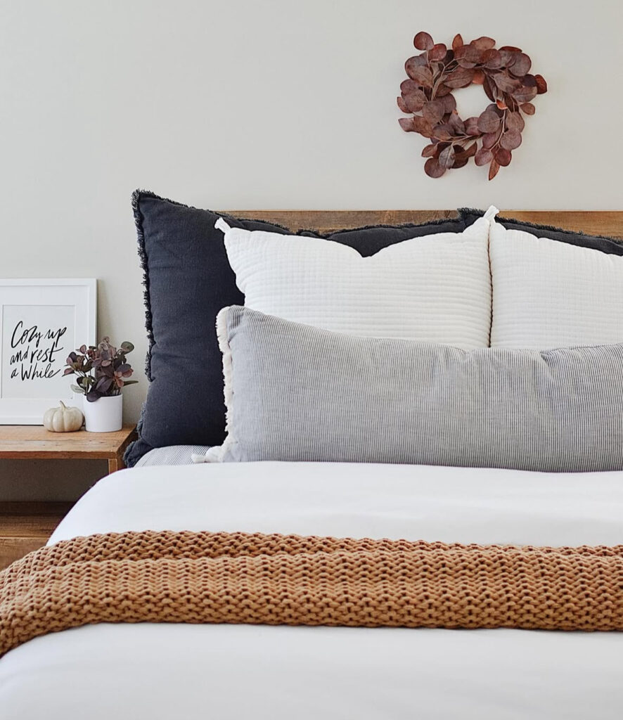 Minimal neutral bed styling with black-and-white pillows, long gray lumbar, tan knit throw and mini pumpkins — fall decor ideas for bedroom.