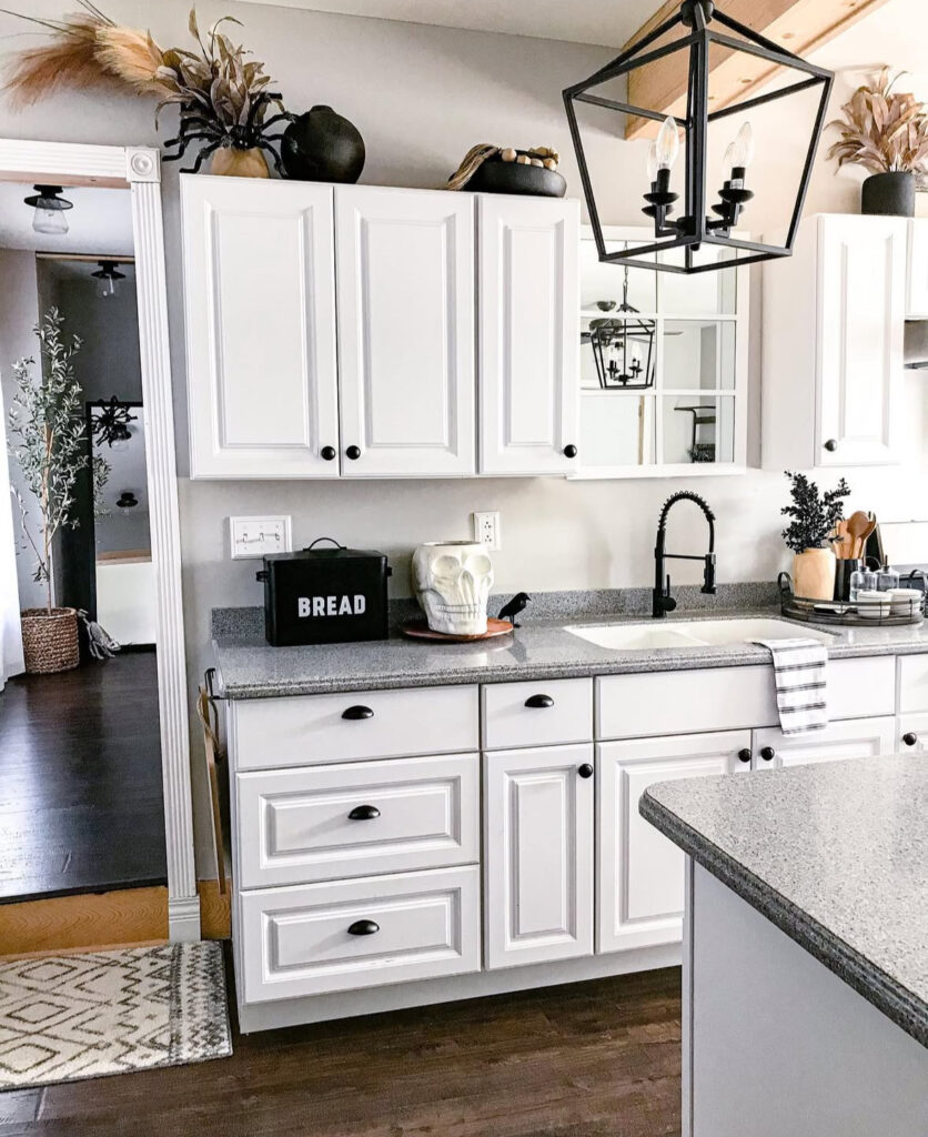 Minimal white-and-wood kitchen with a ceramic skull, matte bread box, and dried grasses above the cabinets.