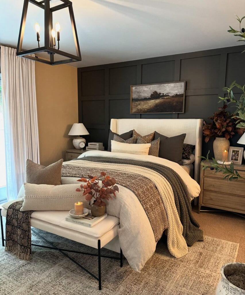Dark paneled wall, layered knits, candlelight and landscape art above upholstered bed — moody fall bedroom decor ideas.