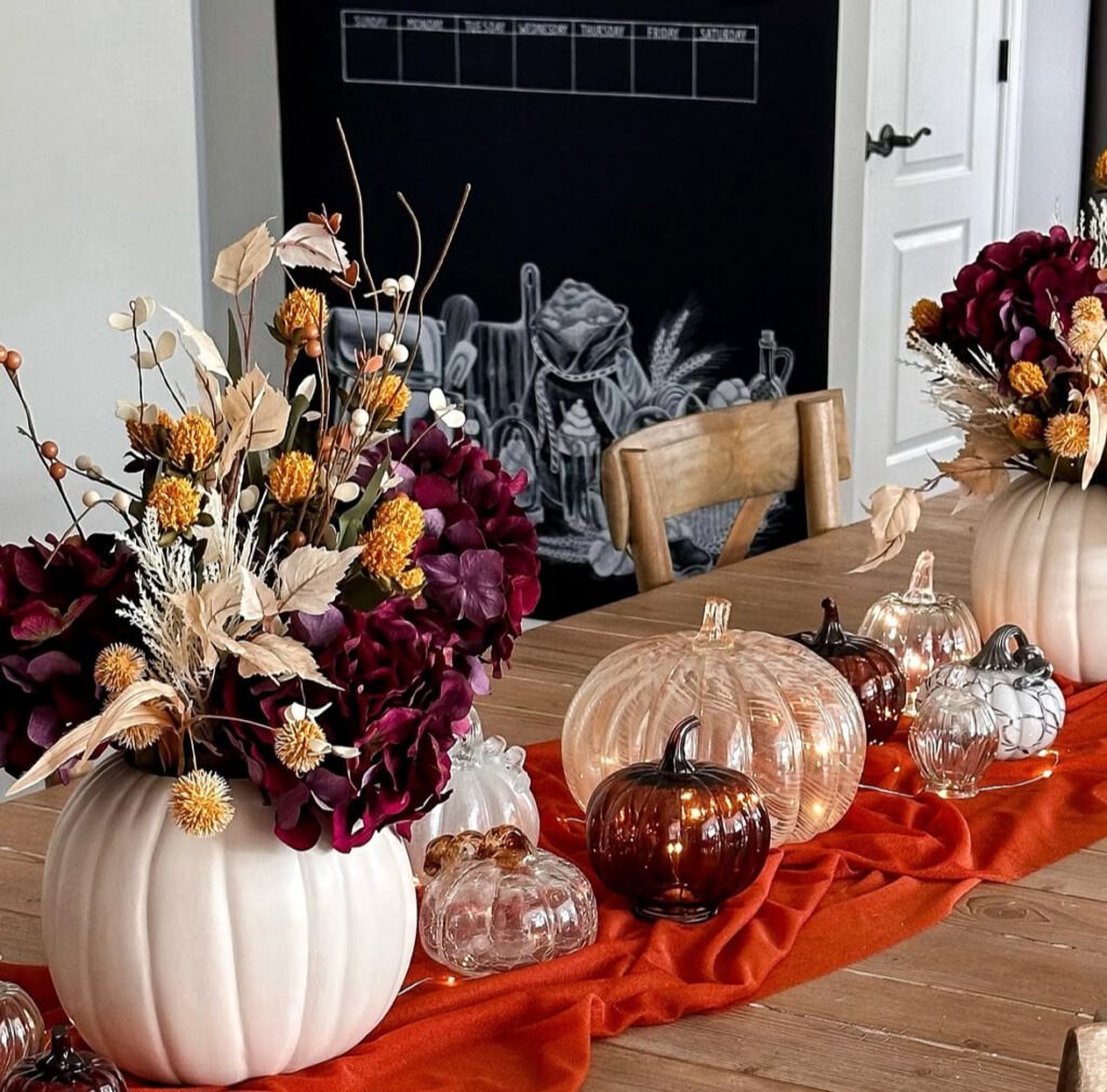 Faux pumpkins used as vases for wine-colored blooms beside glowing glass pumpkins and string lights.