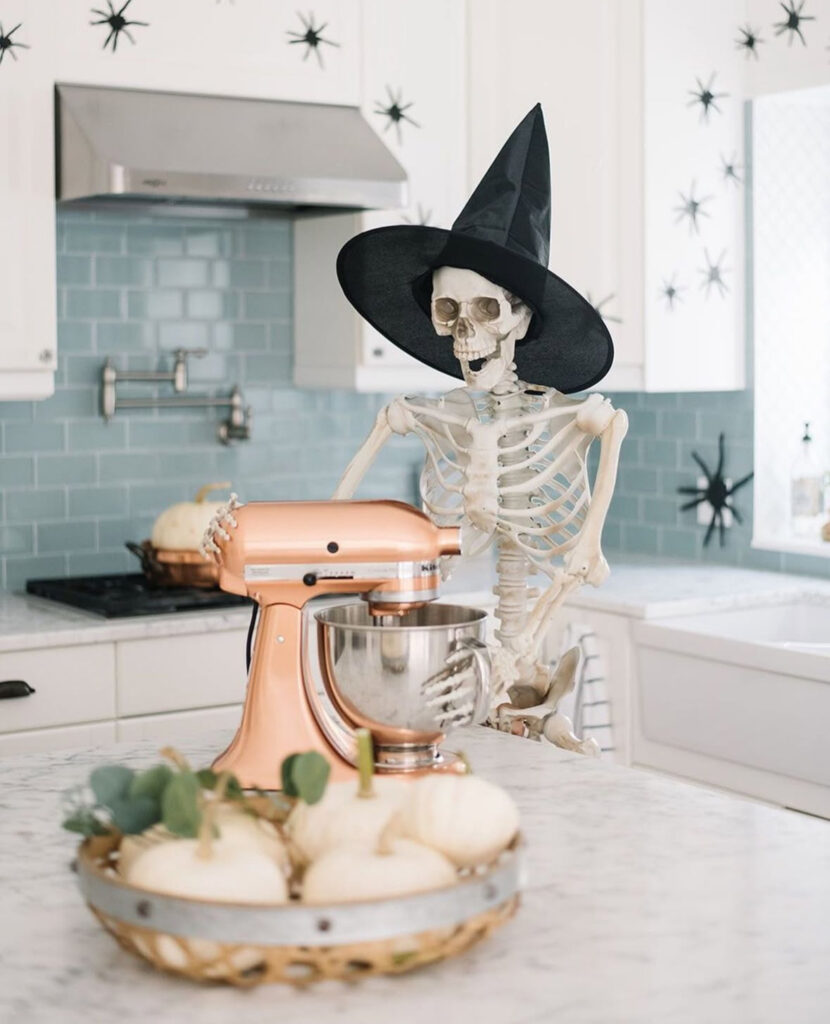 Witch-hat skeleton posed beside a stand mixer with white pumpkins and climbing wall spiders.