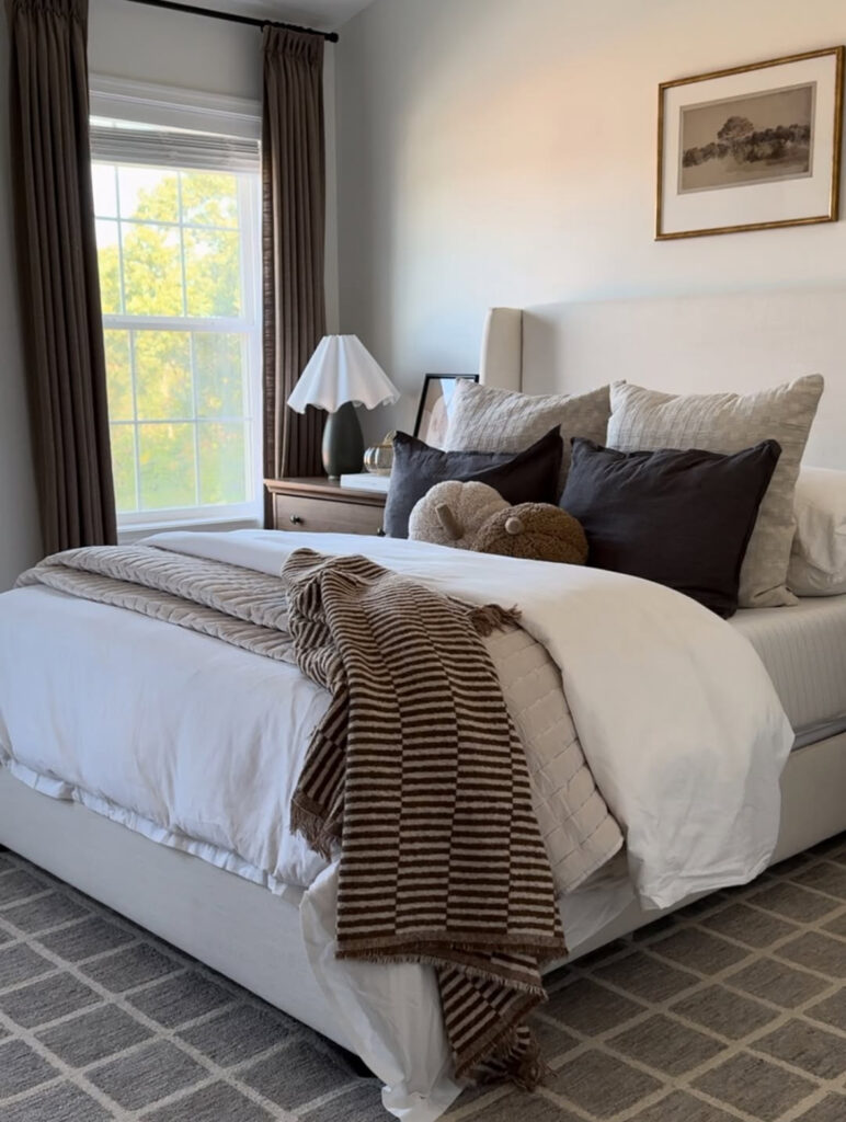 Neutral transitional bedroom with striped brown throw, layered bedding, dark drapes and grid rug — neutral fall bedroom ideas.