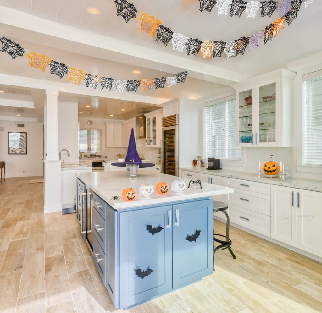 Blue kitchen island with bat decals, ceiling strung with paper spiderweb garlands, and a purple witch hat centerpiece.