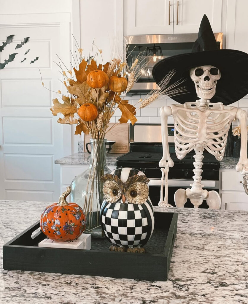 Black tray centerpiece on a granite island with dried stems, floral pumpkin, checkered owl, and a witch-hat skeleton behind.