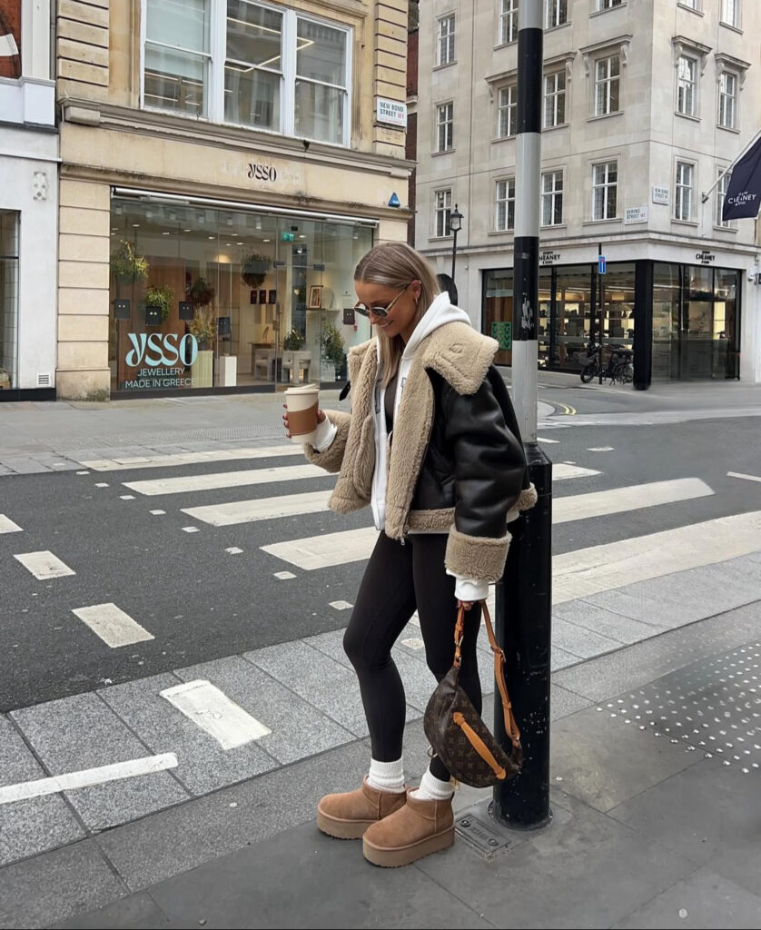 Shearling jacket over hoodie with black leggings and platform mini UGG-style boots; coffee in hand.