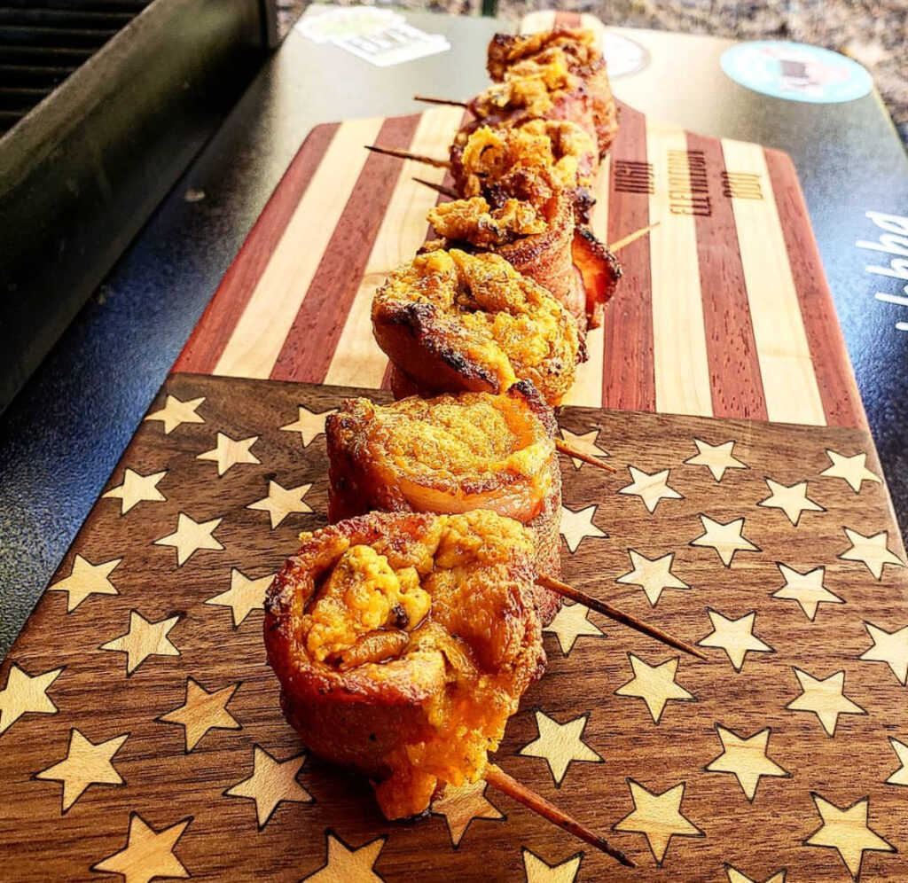 Cylindrical bacon “cups” stuffed with cheesy filling, charred edges, lined on a cutting board.