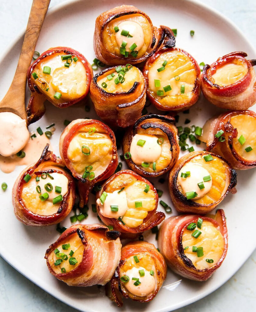 Seared scallops wrapped in bacon with creamy sauce dollops and chopped chives on a white plate.