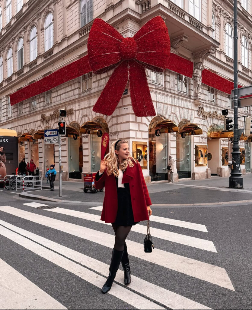 Woman crossing a city street in a red coat, black mini skirt, tights, and knee-high black boots beneath a giant red bow decoration.