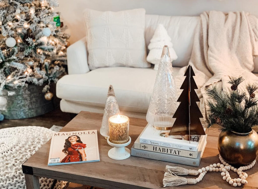 Wood table with glass trees, metal tree silhouette, glowing candle, stacked design books, and a bead tassel; flocked tree in back.