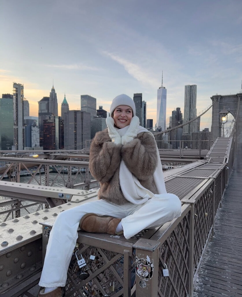 Faux-fur coat, ivory scarf, and cozy boots posing on a bridge with city skyline behind.