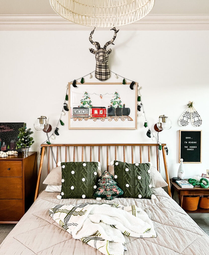 Mid-century bed with green pom pillows, banner, and train art—cute Christmas bedroom ideas in a modern space. Subtle garland and mini tree keep it festive.