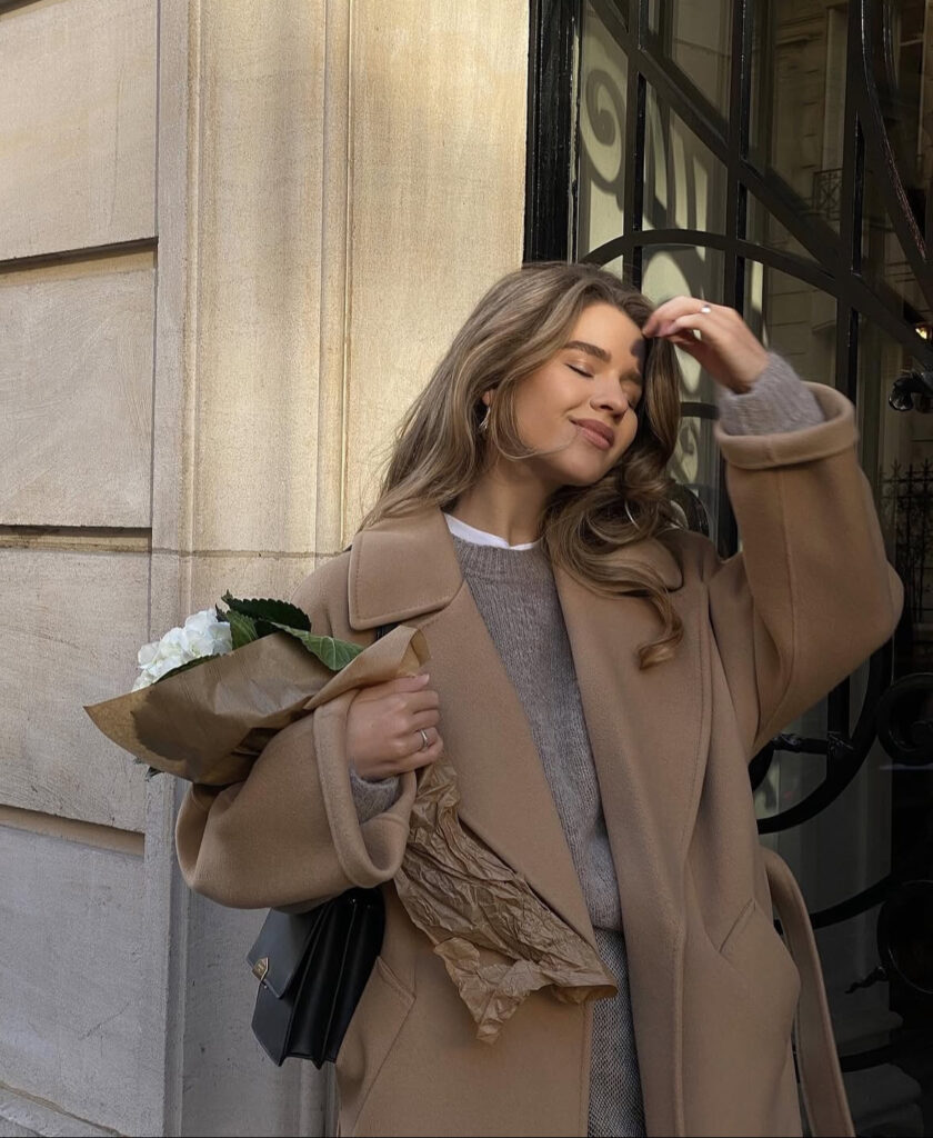 Camel coat layered over knit and white shirt; office-ready look with flowers in hand.
