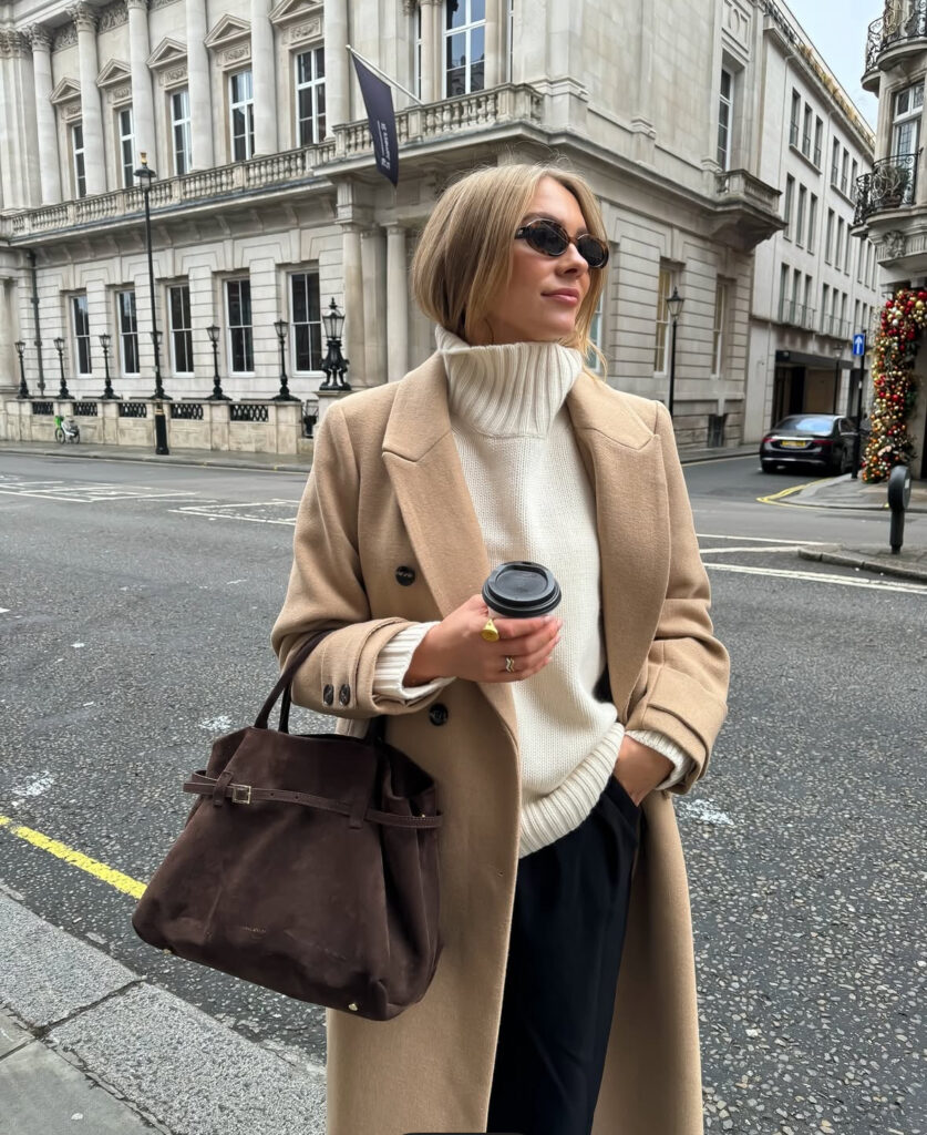 Camel coat over ribbed turtleneck with dark trousers and suede tote; woman holding coffee on a city sidewalk.