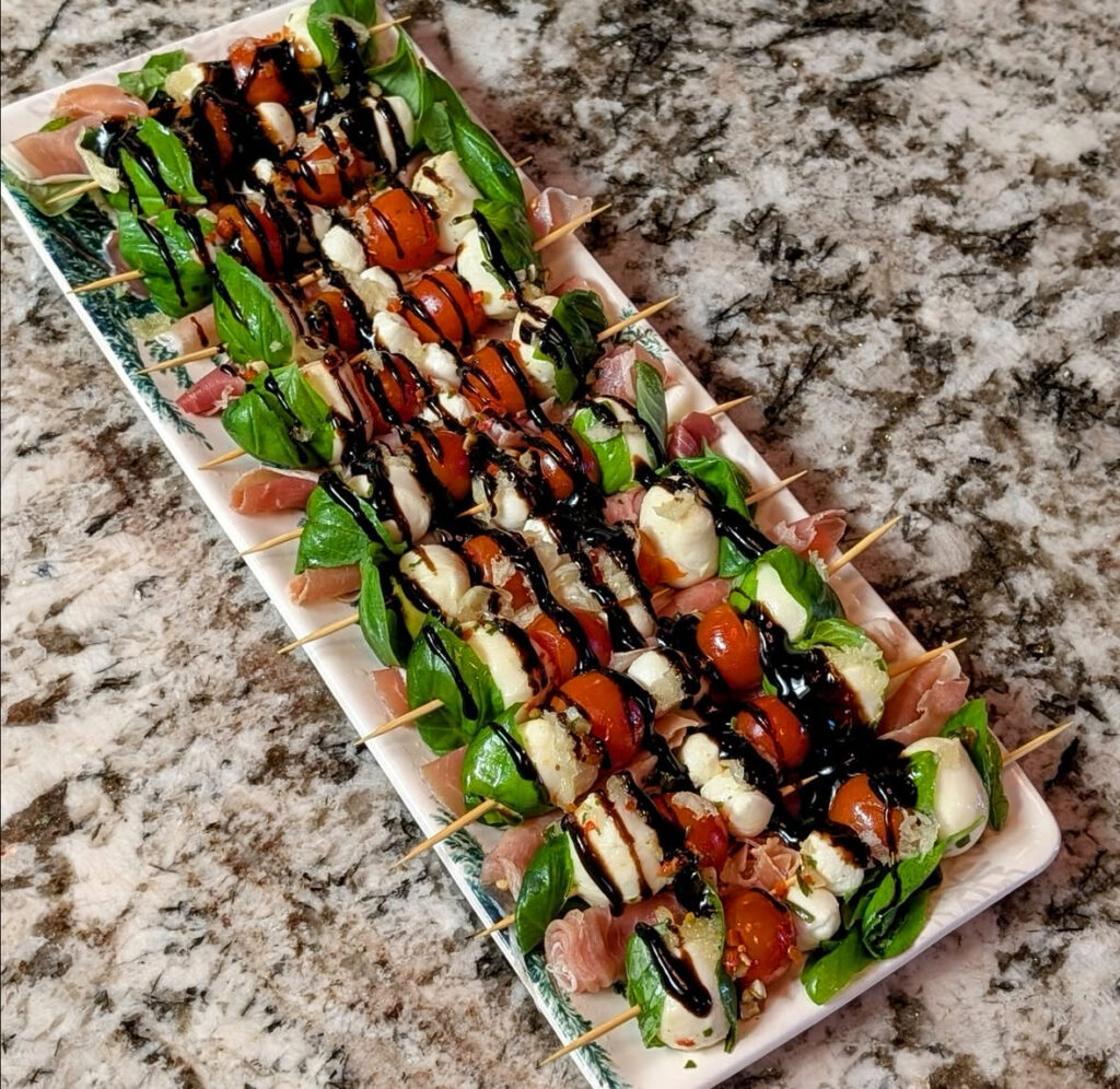 Skewers threaded with basil leaves, cherry tomatoes, mini mozzarella balls, balsamic drizzle, and optional prosciutto.
