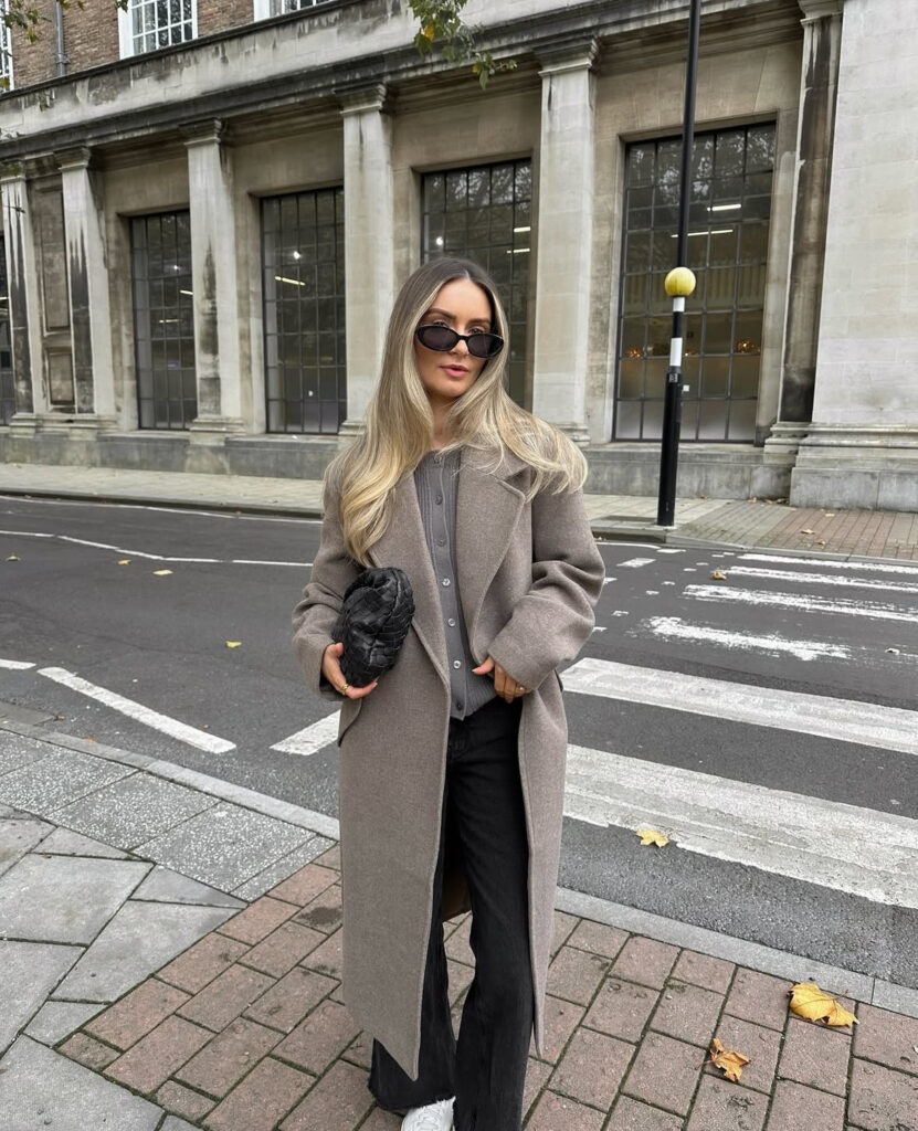 Long tailored coat over cardigan, dark denim, and crisp white sneakers; street-to-desk look.