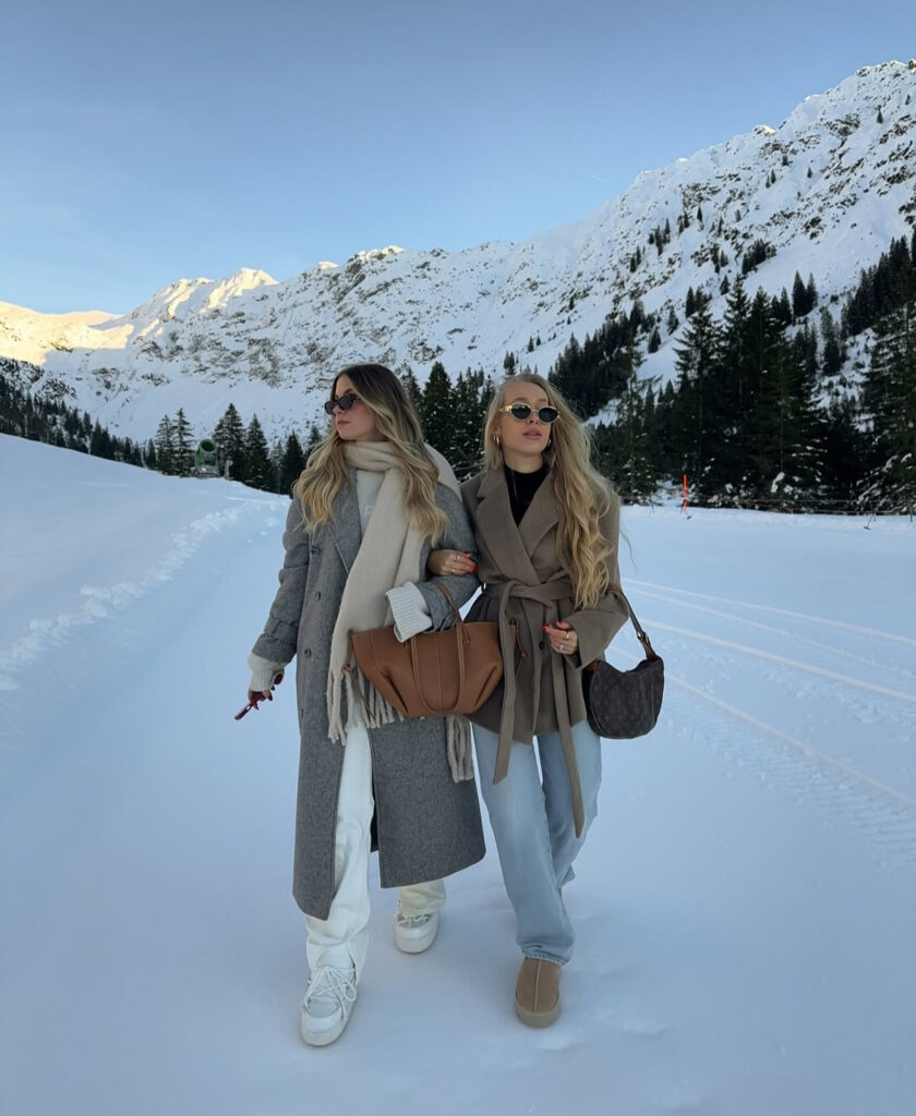 Two friends in coordinated winter neutrals—one in grey and cream, one in taupe and denim—both in sunglasses.