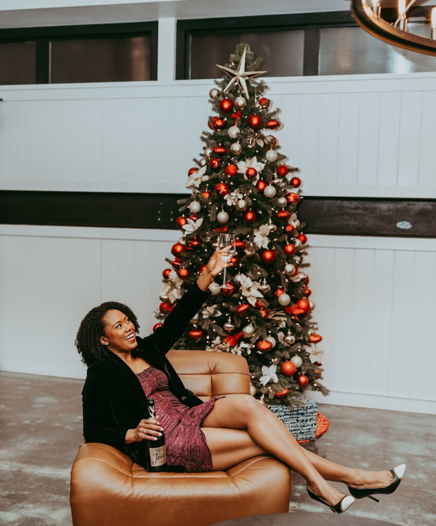 Woman in magenta strapless mini dress with black blazer and slingback heels, raising a champagne glass beside a decorated Christmas tree.