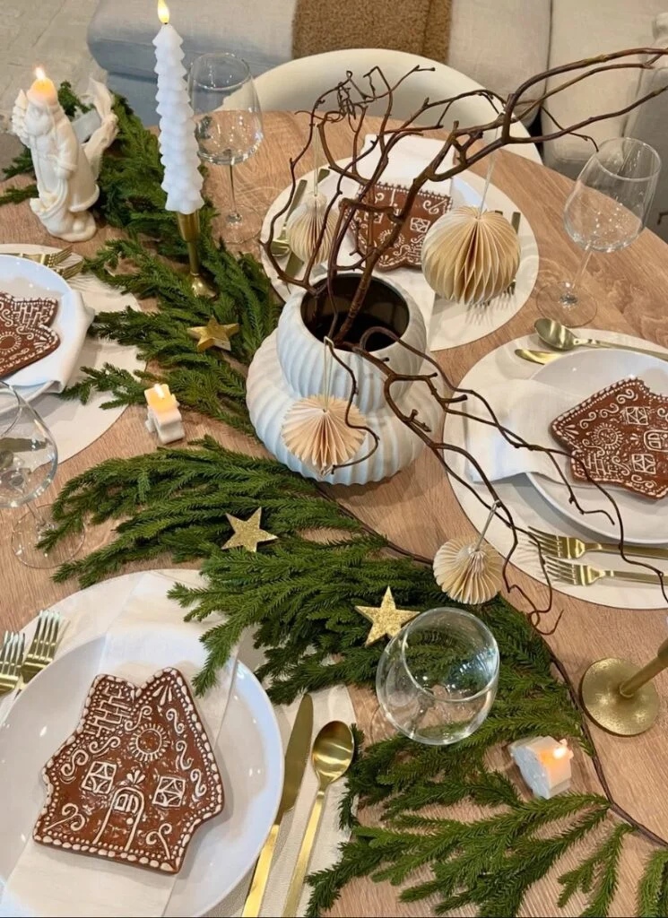Round table styled with cedar garland, gold flatware, sculptural vase of curly branches and paper ornaments, and white plates topped with gingerbread house cookies.