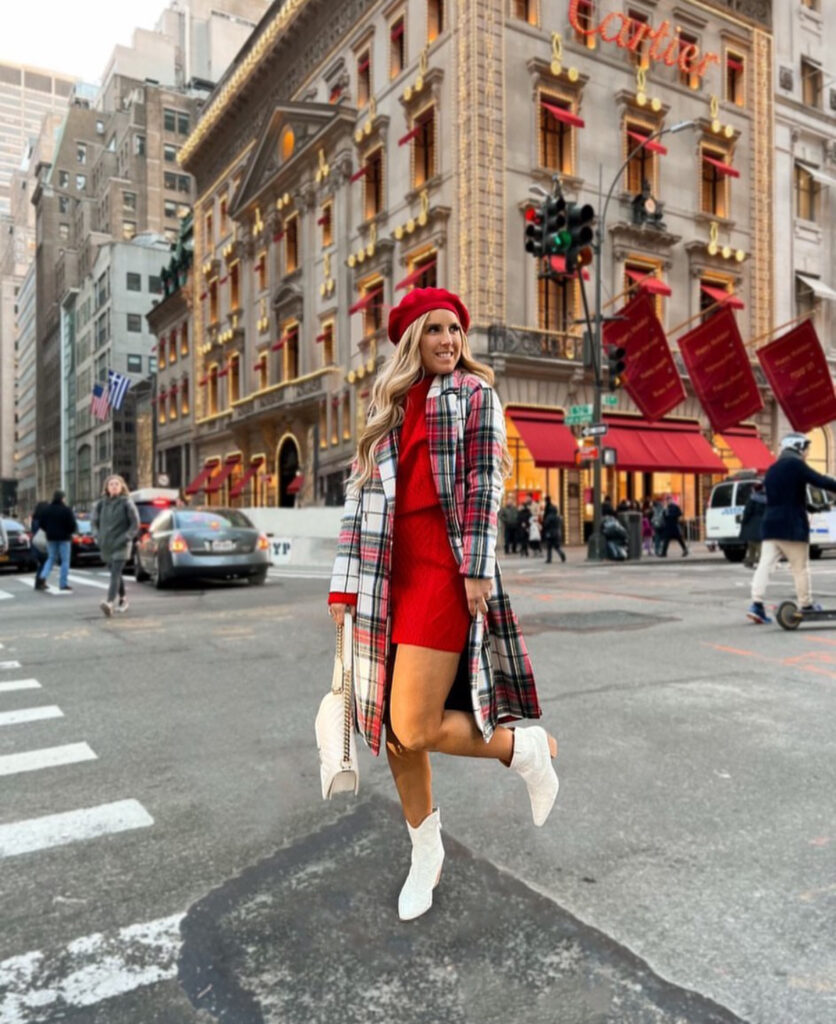 Woman mid-jump on a city street wearing a red knit dress, plaid long coat, red beret, white booties and white bag near Cartier building.