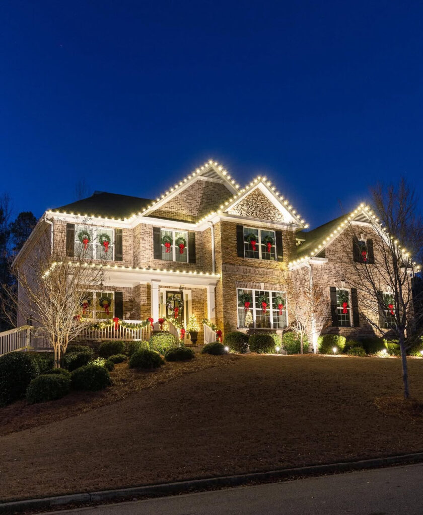 Large brick home with sharp roofline lights and wreaths tied with red bows.