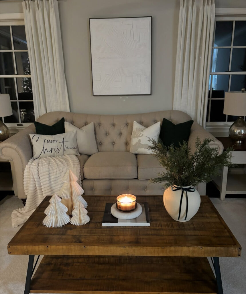 Rustic wood table with paper trees, glowing candle, and white vase of greenery tied with black ribbon in front of a tufted sofa.