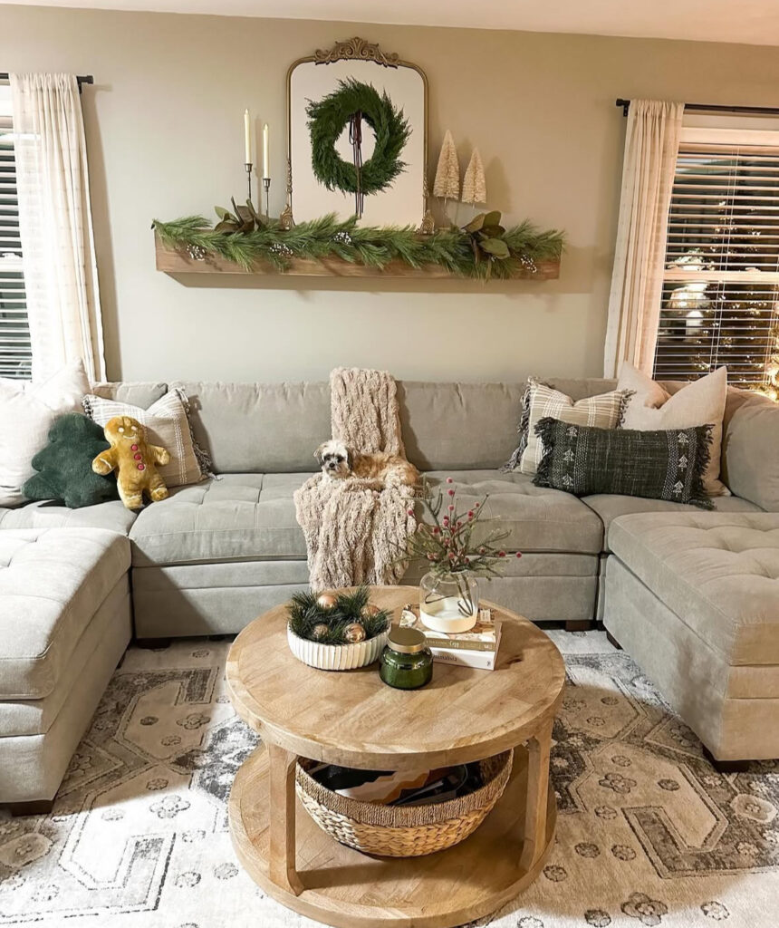 Round wood two-tier table with bowl of greens and ornaments, small vase of berries, candle, and book stack; sectional and shelf with wreath.