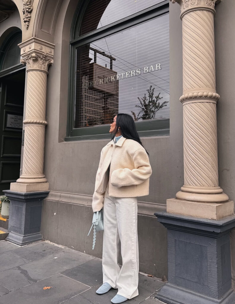 Cream sherpa jacket with wide-leg ecru jeans, pale blue bag and flats on a bright winter day.