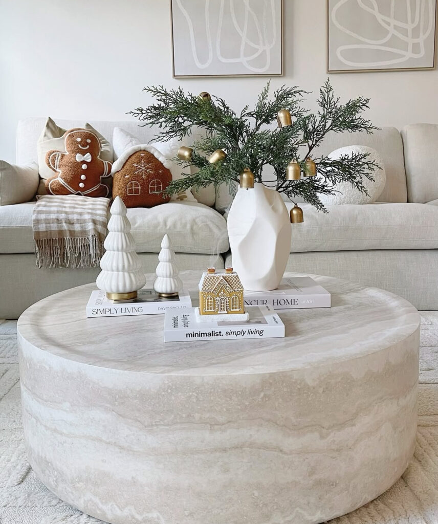Large travertine round coffee table with sculptural white vase of greens and brass bells, ceramic trees, and a mini gingerbread house.