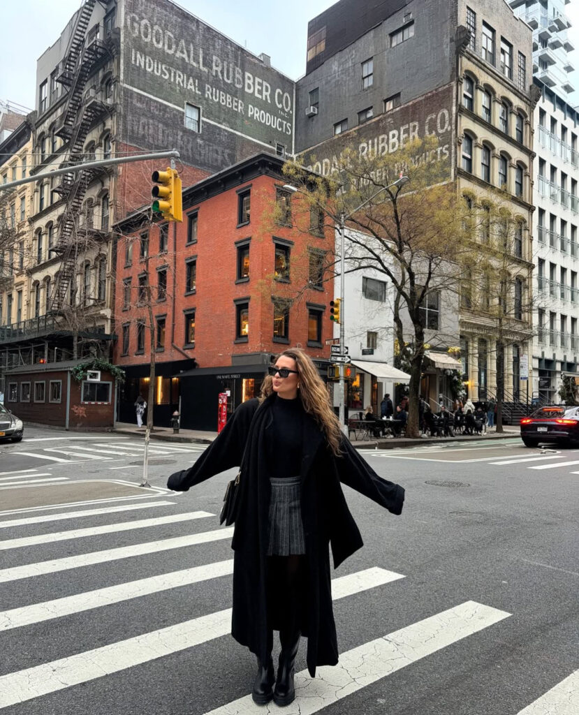 Long black coat, knit turtleneck, knee-boots, and pleated mini skirt on a downtown crosswalk.