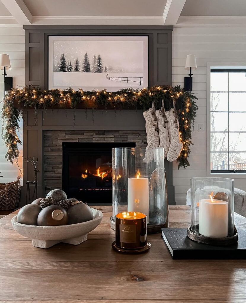 Wood table in front of a fireplace; hurricanes with pillar candles, amber jar candle, and a stone bowl of matte ornaments.