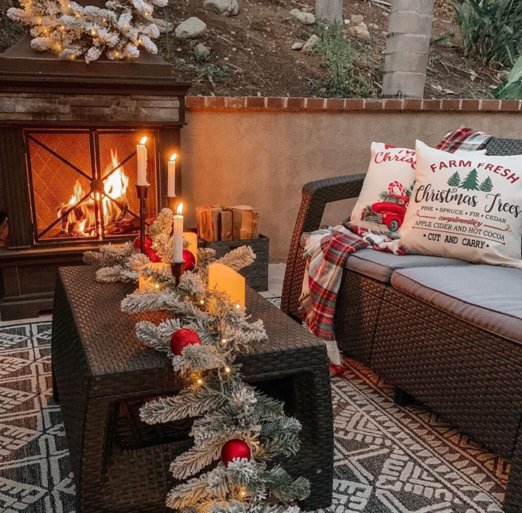 Outdoor fireplace patio with candles, flocked garland, plaid throw, and Christmas pillows.