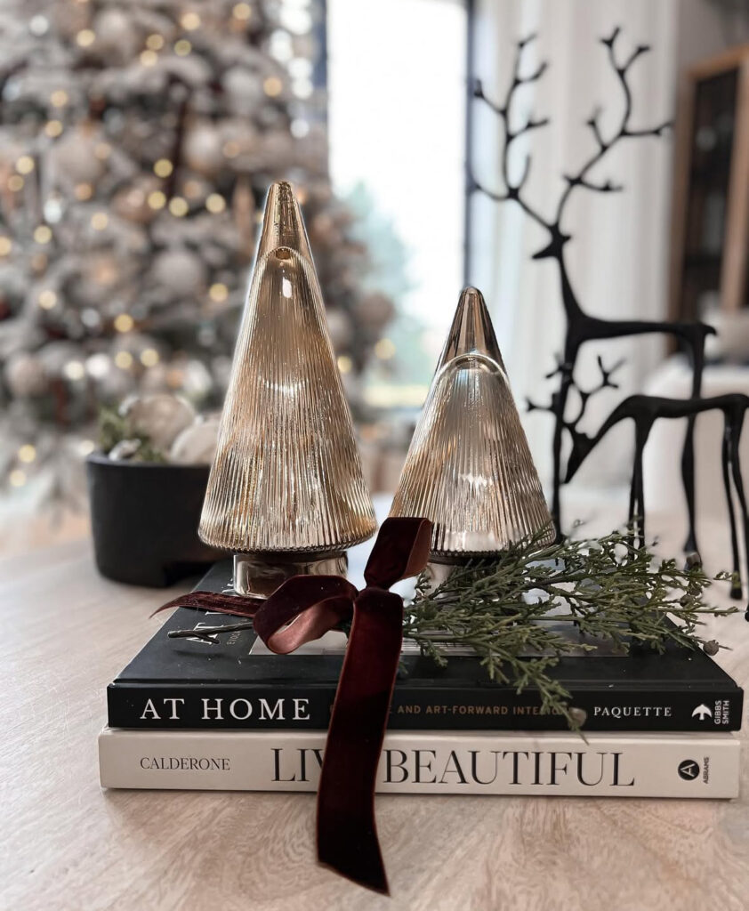 Two mercury-glass trees displayed on stacked coffee-table books with a velvet ribbon and a small greenery sprig; blurred reindeer behind.