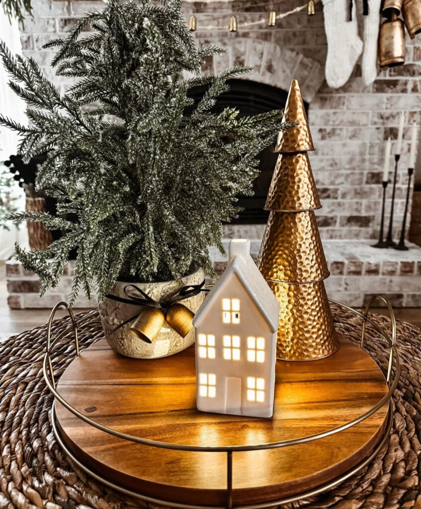 Round tray with a lit ceramic house, hammered brass tree, and frosted potted greenery tied with jingle bells.