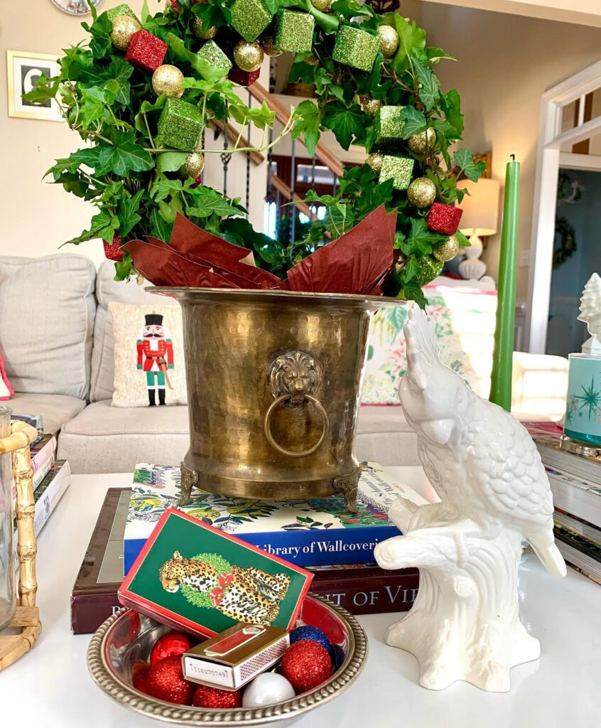 Vintage brass champagne bucket with ivy wreath and glitter cubes on stacked books; white parrot figurine and bowl of ornaments with matches.
