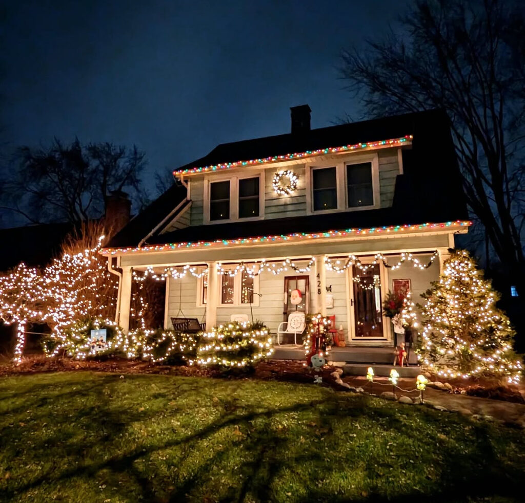 Classic house outlined with warm roofline lights, wreaths in windows, glowing shrubs.