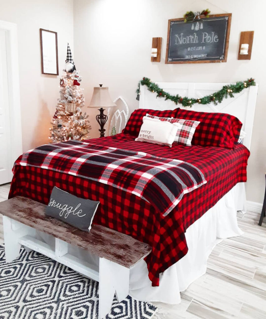 Buffalo-check bedding with chalkboard “North Pole” sign—rustic cozy Christmas bedroom ideas. A tiny corner tree and bench invite snuggling.