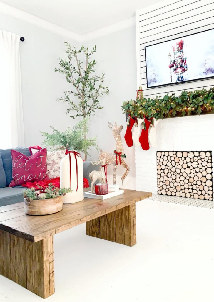 Rustic plank coffee table with large pillar “vase” holding greenery, bowl of evergreens, red candle, and wicker reindeer; stockings on mantle.