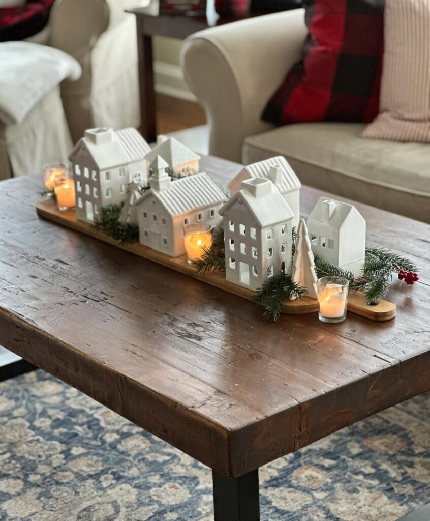 Long wood tray filled with white ceramic village houses, tealights, and fresh evergreen sprigs on a dark coffee table.