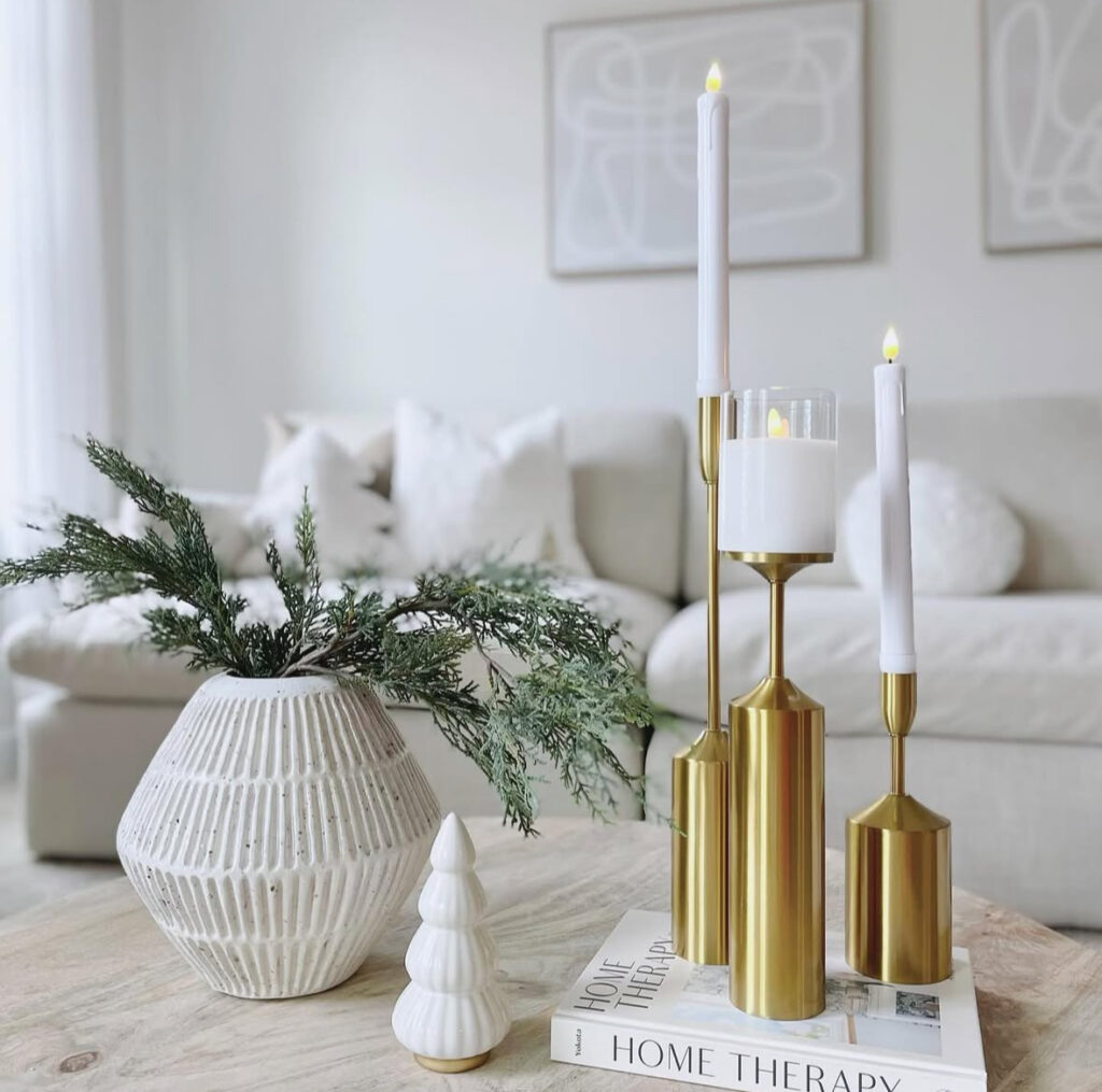 Brass candlesticks with white tapers, ribbed white vase of greenery, and a small ceramic tree styled on books on a light coffee table in a bright neutral living room.