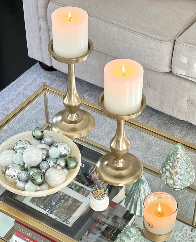 Brass candleholders on a glass table beside a marble bowl of mint and silver ornaments, small glass trees, and a scented candle.