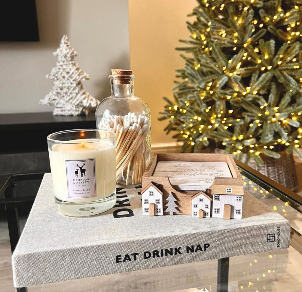 Minimal glass candle, jar of matches, and wooden house coasters styled on a linen book on a glass coffee table with lit tree in background.