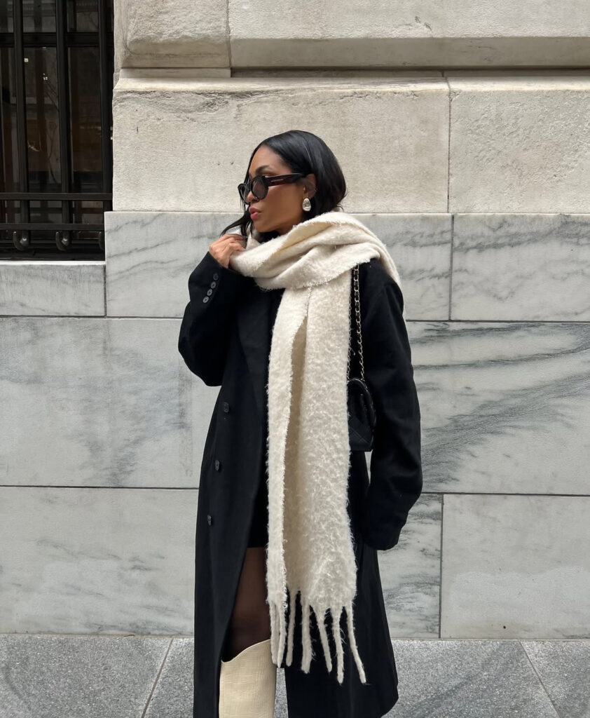 Long black coat with dramatic ivory scarf and knee-high boots; sleek monochrome street style.
