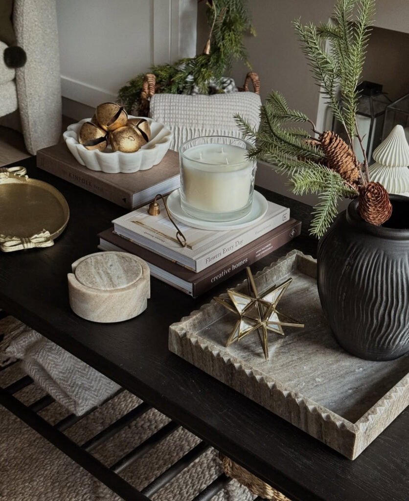 Dark wood table layered with books, a marble coaster box, large candle, black vase of evergreens and pinecone, scalloped bowl of jingle bells, and a stone tray with brass star.