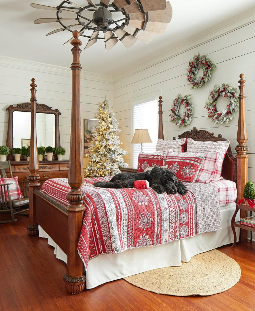 Carved four-poster bed with sweater-knit linens and trio of wreaths—classic Christmas bedroom decor ideas. Flocked tree glows in the corner.